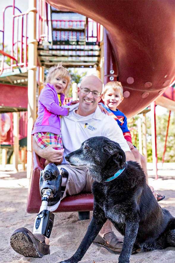 Man smiling while sitting on slide with kids and dog with a prosthetic leg 
