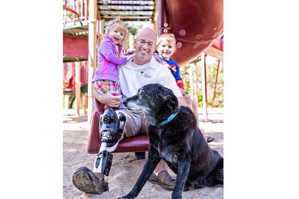 Ben Blecha sitting on slide, wearing a prosthetic, with his kids on his lap and dog sitting in front of him